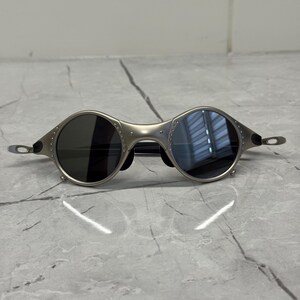 May include: Silver-toned sunglasses with round lenses. The frames have a unique, industrial design with visible rivets. The lenses are dark, providing a contrast to the metallic frame. The temples are thin and extend to the ear pieces.