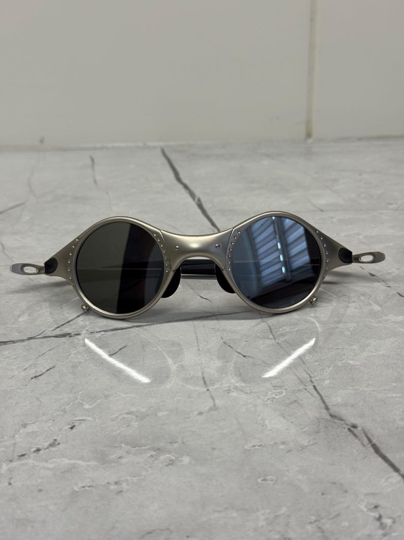 May include: Silver-toned sunglasses with round lenses. The frames have a unique, industrial design with visible rivets. The lenses are dark, providing a contrast to the metallic frame. The temples are thin and extend to the ear pieces.
