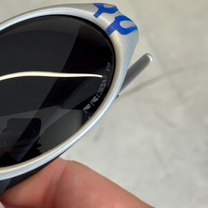 Puede incluir: Gafas de sol plateadas con lentes oscuras. Las patillas presentan un dise&ntilde;o azul y plateado. Las lentes tienen una ligera curvatura y la montura parece ser de metal. Unas manos sostienen las gafas.
