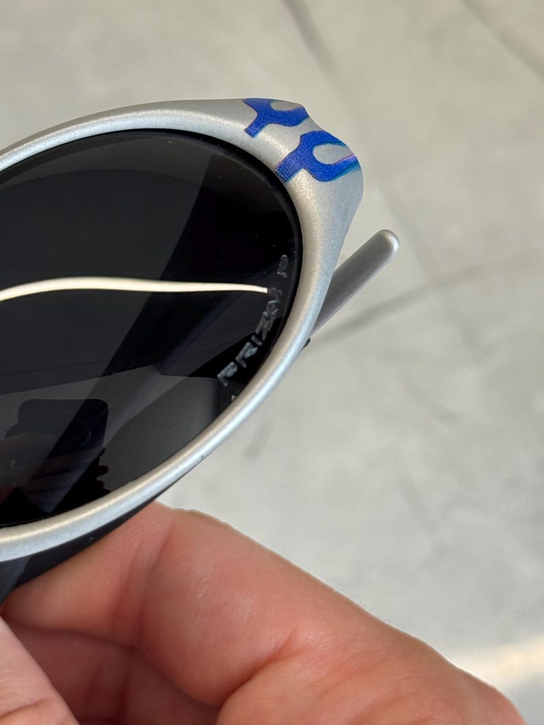 Puede incluir: Gafas de sol plateadas con lentes oscuras. Las patillas presentan un dise&ntilde;o azul y plateado. Las lentes tienen una ligera curvatura y la montura parece ser de metal. Unas manos sostienen las gafas.