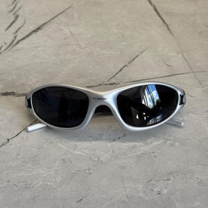May include: Silver-framed sunglasses with dark lenses. The sunglasses have a sleek, sporty design with a curved frame and a small brand name on the top of the frame. The lenses reflect the surrounding environment.