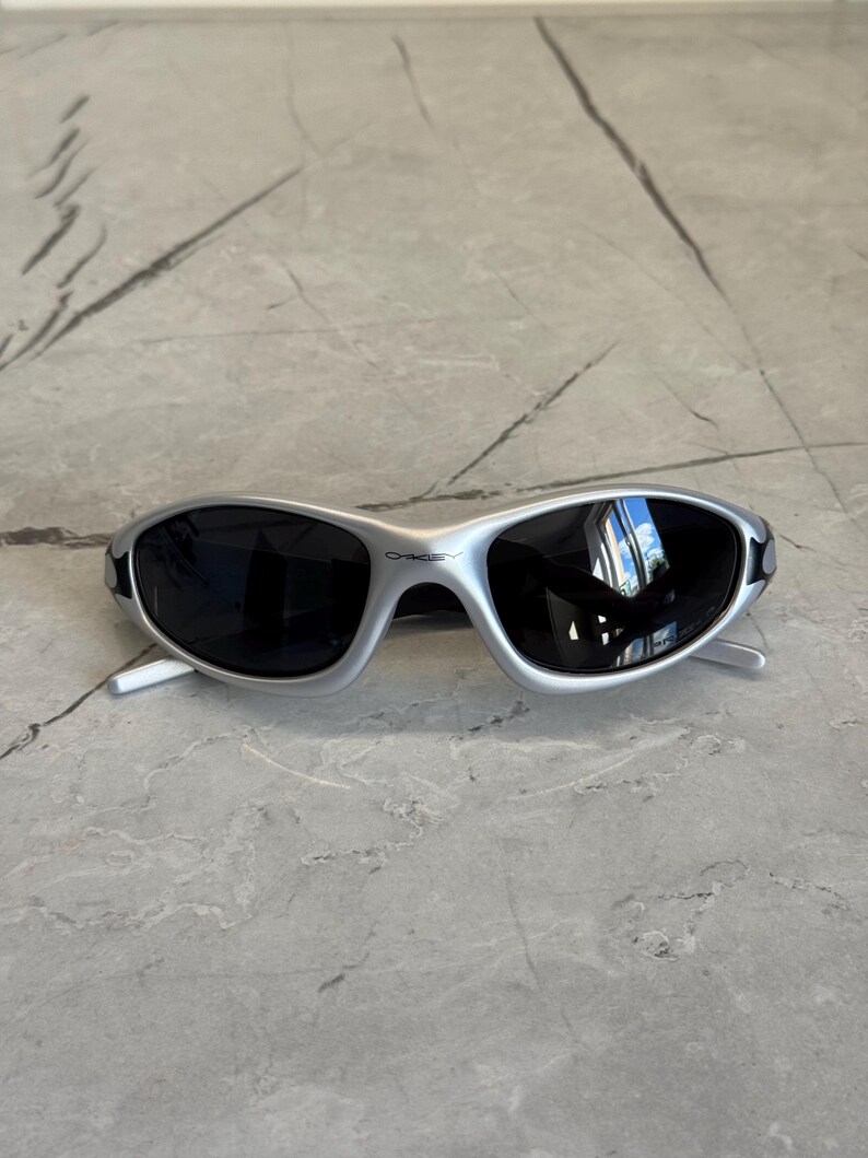 May include: Silver-framed sunglasses with dark lenses. The sunglasses have a sleek, sporty design with a curved frame and a small brand name on the top of the frame. The lenses reflect the surrounding environment.
