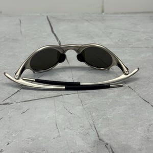 Może przedstawiać: Okulary przeciwsłoneczne w srebrnej oprawie z ciemnymi soczewkami. Okulary przeciwsłoneczne mają elegancki, nowoczesny design z zakrzywioną oprawą i czarnym gumowym noskiem. Zauszniki są cienkie i rozciągają się na zewnątrz.