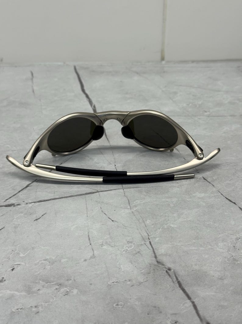 Może przedstawiać: Okulary przeciwsłoneczne w srebrnej oprawie z ciemnymi soczewkami. Okulary przeciwsłoneczne mają elegancki, nowoczesny design z zakrzywioną oprawą i czarnym gumowym noskiem. Zauszniki są cienkie i rozciągają się na zewnątrz.