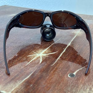 Puede incluir: Gafas de sol negras y marrones con lentes oscuras. Las gafas de sol est&aacute;n sobre una superficie de madera. Un peque&ntilde;o objeto cil&iacute;ndrico negro est&aacute; posicionado entre las lentes.