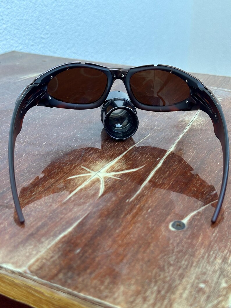 Puede incluir: Gafas de sol negras y marrones con lentes oscuras. Las gafas de sol est&aacute;n sobre una superficie de madera. Un peque&ntilde;o objeto cil&iacute;ndrico negro est&aacute; posicionado entre las lentes.