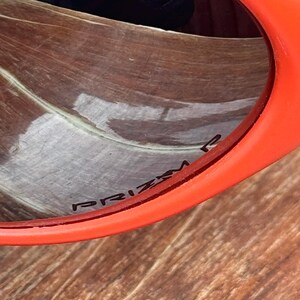 Pu&ograve; includere: Primo piano di occhiali da sole arancioni con la scritta "PRIZM" stampata sulla lente. Gli occhiali da sole hanno una montatura spessa e arrotondata. Lo sfondo &egrave; una superficie di legno.