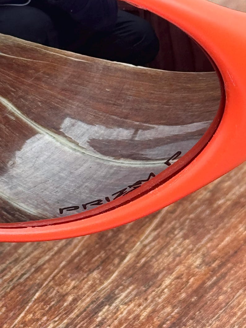 Pu&ograve; includere: Primo piano di occhiali da sole arancioni con la scritta "PRIZM" stampata sulla lente. Gli occhiali da sole hanno una montatura spessa e arrotondata. Lo sfondo &egrave; una superficie di legno.
