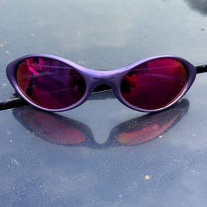 May include: Purple sunglasses with a curved frame and reflective red lenses. The sunglasses have a small bridge and a subtle, rounded shape. The lenses reflect the surrounding environment, creating a mirrored effect.