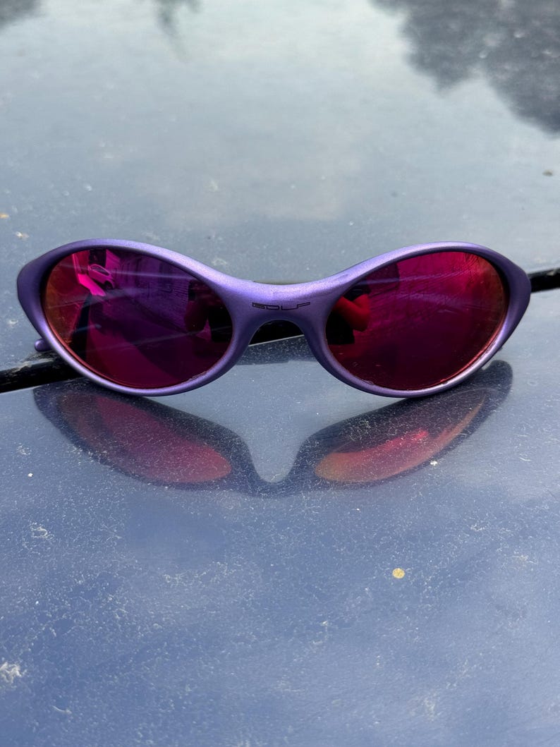 May include: Purple sunglasses with a curved frame and reflective red lenses. The sunglasses have a small bridge and a subtle, rounded shape. The lenses reflect the surrounding environment, creating a mirrored effect.