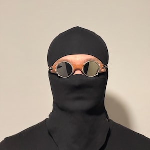 May include: A person wearing a black balaclava and a black face covering. The person is wearing round, silver-rimmed sunglasses with mirrored lenses. The background is a solid, neutral color.