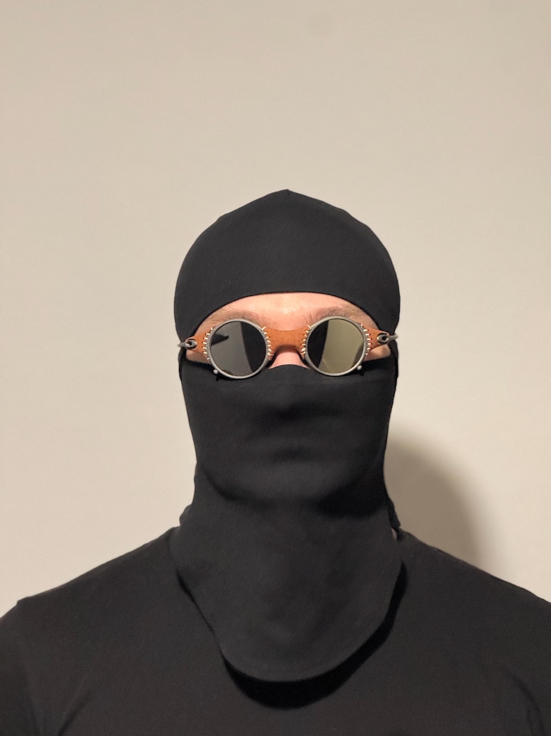 May include: A person wearing a black balaclava and a black face covering. The person is wearing round, silver-rimmed sunglasses with mirrored lenses. The background is a solid, neutral color.