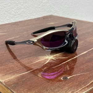 May include: A pair of silver and black sunglasses with purple lenses. The sunglasses are sitting on a wooden surface.