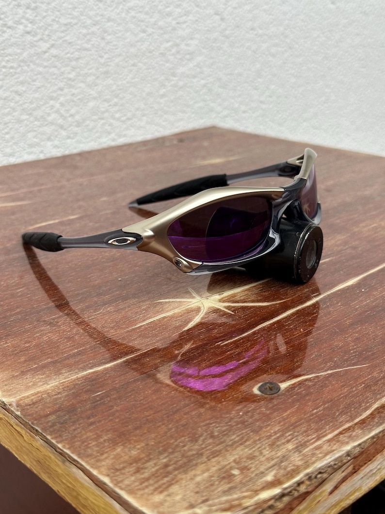 May include: A pair of silver and black sunglasses with purple lenses. The sunglasses are sitting on a wooden surface.