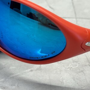 Op de afbeelding: Close-up van een oranje zonnebril met blauwe spiegelglazen. De glazen reflecteren de omgeving. De tekst "PRIZM P" is zichtbaar op de lens. De zonnebril ligt op een lichtgrijze ondergrond.