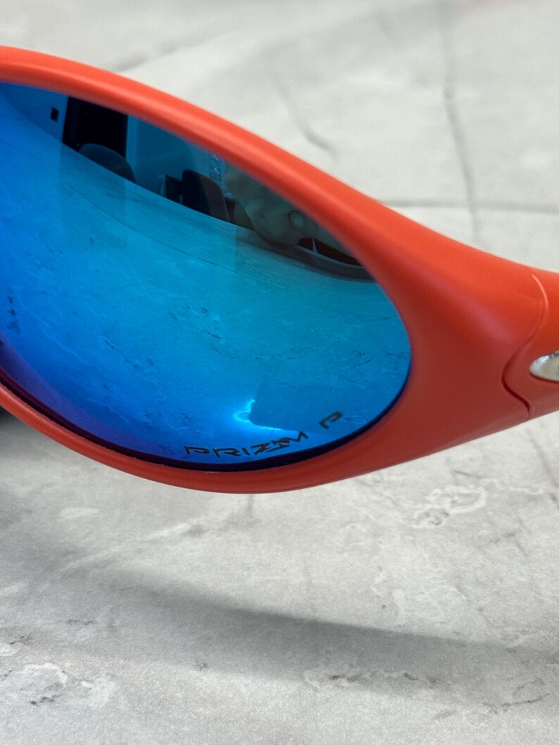 Op de afbeelding: Close-up van een oranje zonnebril met blauwe spiegelglazen. De glazen reflecteren de omgeving. De tekst "PRIZM P" is zichtbaar op de lens. De zonnebril ligt op een lichtgrijze ondergrond.