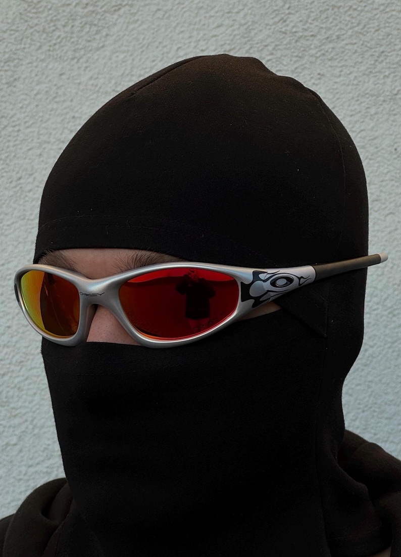 May include: Silver-framed sunglasses with red lenses. The sunglasses have a curved design and are worn with a black balaclava. The lenses reflect the surrounding environment. The sunglasses have a decorative design on the temples.