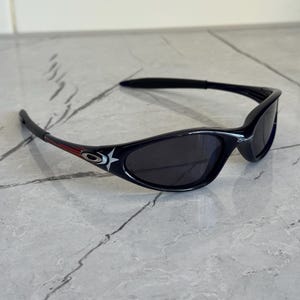 K&ouml;nnte beinhalten: Schwarze Sonnenbrille mit geschwungenem Rahmen und dunklen Gl&auml;sern. Die B&uuml;gel haben einen roten Streifen und ein wei&szlig;es Sternlogo. Die Sonnenbrille ist f&uuml;r Sport und Outdoor-Aktivit&auml;ten konzipiert.