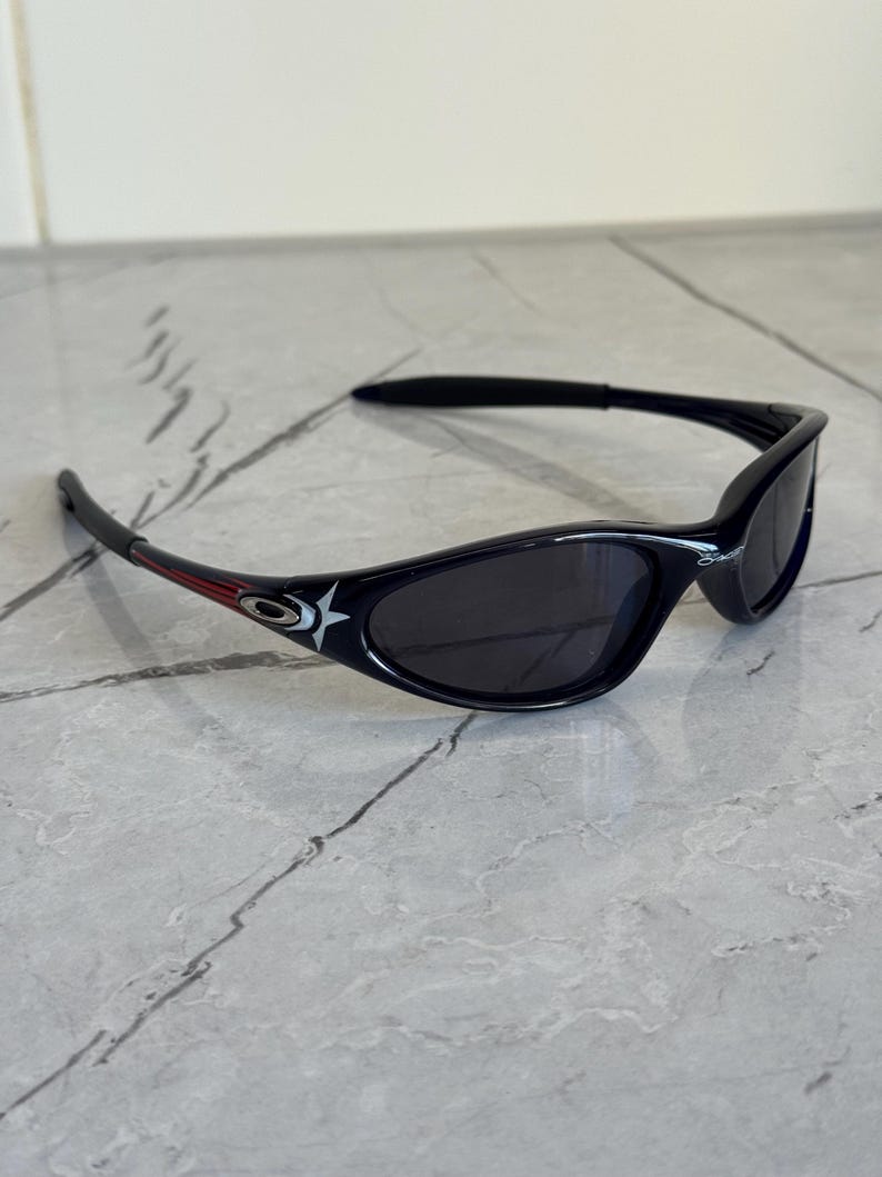 K&ouml;nnte beinhalten: Schwarze Sonnenbrille mit geschwungenem Rahmen und dunklen Gl&auml;sern. Die B&uuml;gel haben einen roten Streifen und ein wei&szlig;es Sternlogo. Die Sonnenbrille ist f&uuml;r Sport und Outdoor-Aktivit&auml;ten konzipiert.
