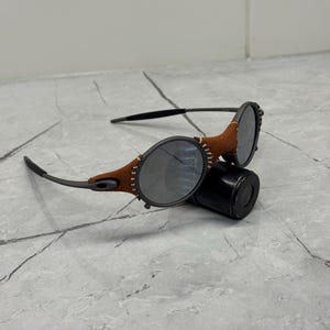 May include: A pair of sunglasses with oval lenses and a brown leather accent around the frames. The temples are black, and the lenses are a dark gray color. The sunglasses are resting on a black cylindrical case.