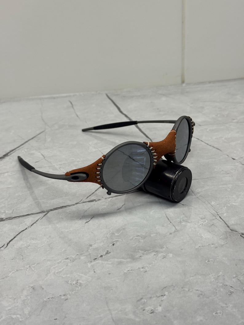 May include: A pair of sunglasses with oval lenses and a brown leather accent around the frames. The temples are black, and the lenses are a dark gray color. The sunglasses are resting on a black cylindrical case.