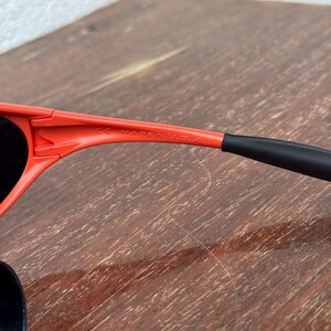 Pu&ograve; includere: Primo piano di occhiali da sole rossi con lenti nere e aste nere. Il testo "MADE IN U.S.A." &egrave; visibile sulla montatura. Gli occhiali da sole sono su una superficie di legno.