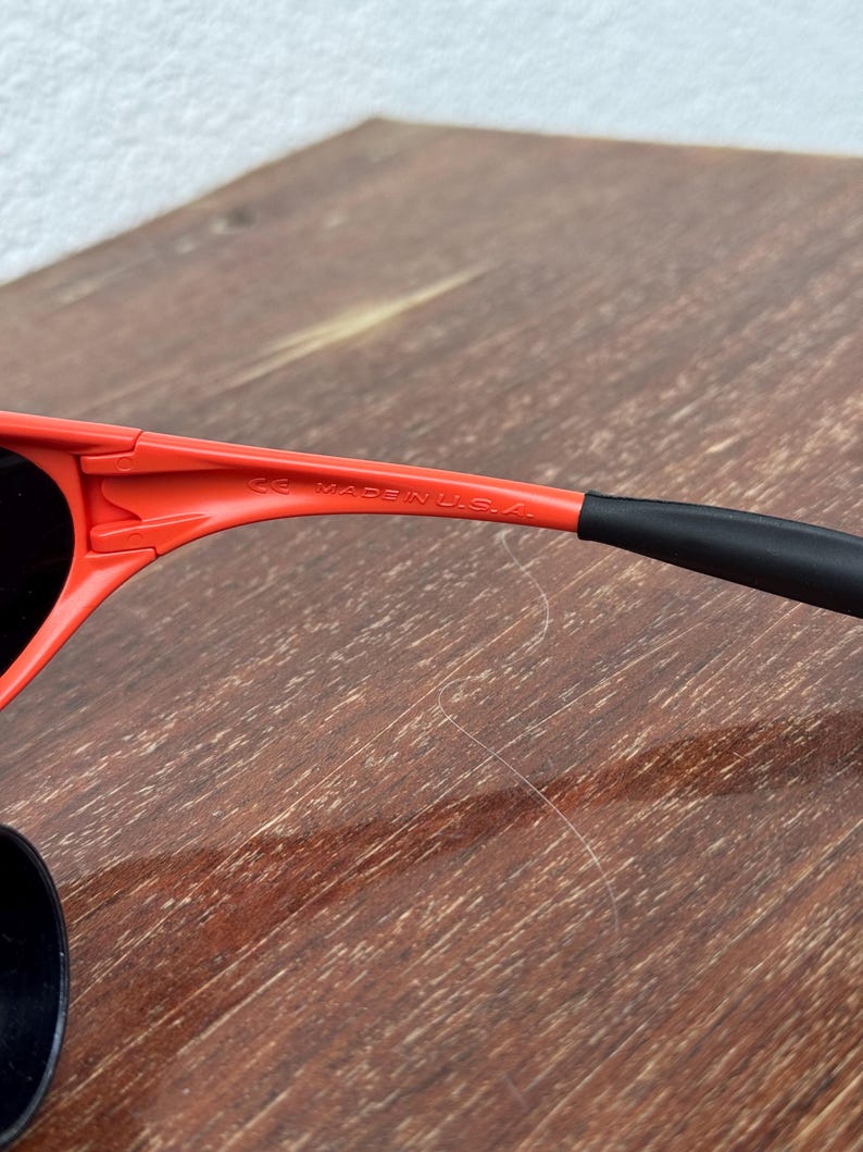 Pu&ograve; includere: Primo piano di occhiali da sole rossi con lenti nere e aste nere. Il testo "MADE IN U.S.A." &egrave; visibile sulla montatura. Gli occhiali da sole sono su una superficie di legno.