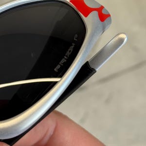 May include: Silver sunglasses with black lenses and red and gray flame accents on the frame. The text "PRIZM P" is visible on the lens. The sunglasses have a sleek, modern design.