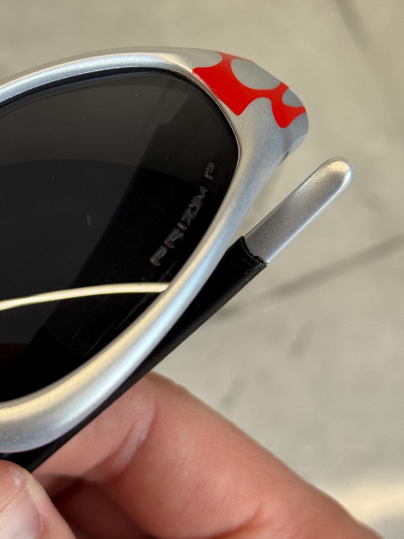 May include: Silver sunglasses with black lenses and red and gray flame accents on the frame. The text "PRIZM P" is visible on the lens. The sunglasses have a sleek, modern design.