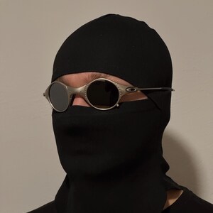 May include: Oval-shaped sunglasses with a silver frame and dark lenses. The sunglasses are worn over a black balaclava. The balaclava covers the head and neck, with an opening for the eyes.