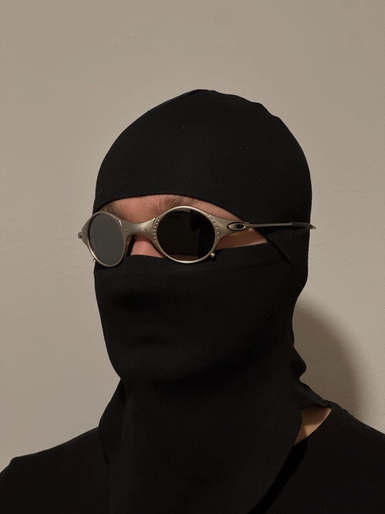 May include: Oval-shaped sunglasses with a silver frame and dark lenses. The sunglasses are worn over a black balaclava. The balaclava covers the head and neck, with an opening for the eyes.