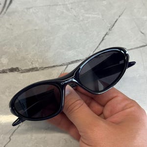K&ouml;nnte beinhalten: Schwarze ovale Sonnenbrille mit dunklen Gl&auml;sern. Die Sonnenbrille hat ein schlankes, modernes Design mit einem d&uuml;nnen Rahmen und dem Markennamen "Oakley" in Wei&szlig; auf dem B&uuml;gel. Die Sonnenbrille wird in einer Hand gehalten.