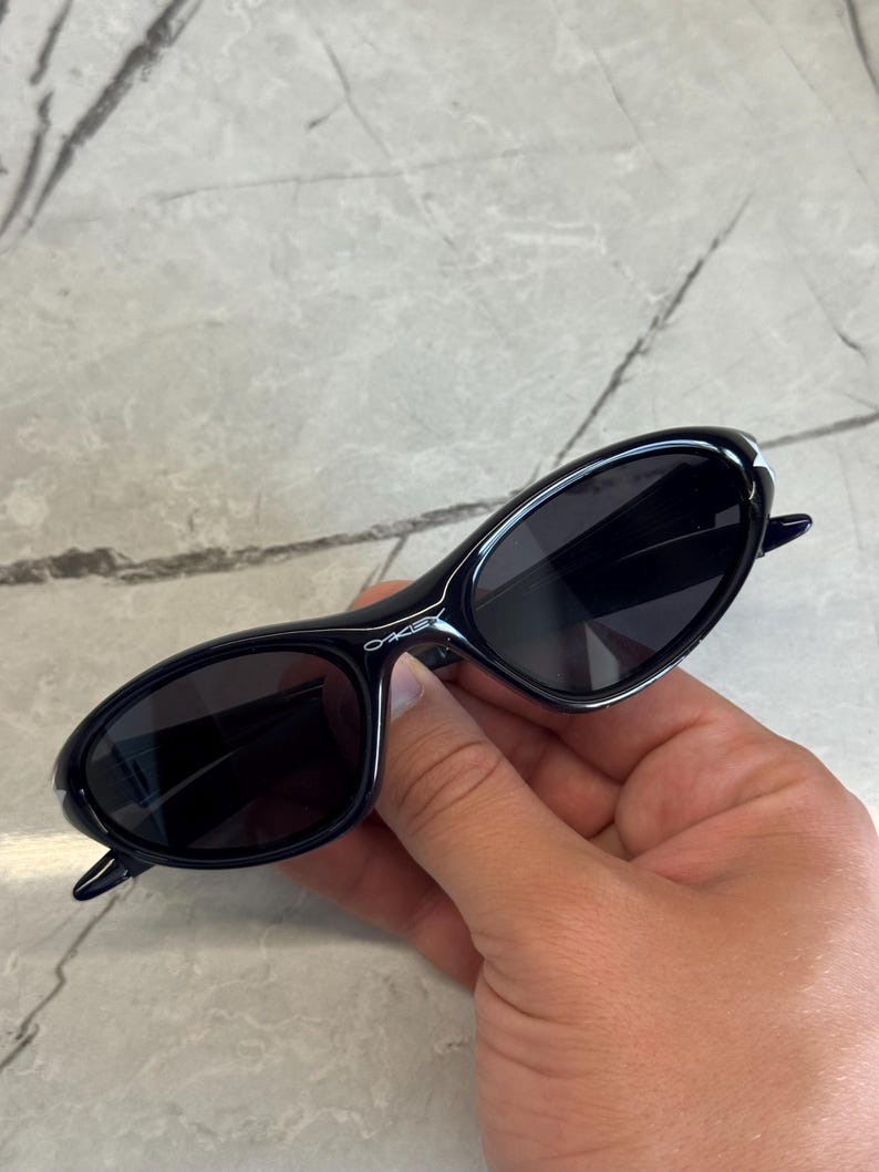 K&ouml;nnte beinhalten: Schwarze ovale Sonnenbrille mit dunklen Gl&auml;sern. Die Sonnenbrille hat ein schlankes, modernes Design mit einem d&uuml;nnen Rahmen und dem Markennamen "Oakley" in Wei&szlig; auf dem B&uuml;gel. Die Sonnenbrille wird in einer Hand gehalten.