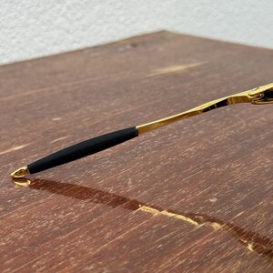 May include: Close-up of a gold-colored eyeglass arm with a black rubber grip. The arm is resting on a wooden surface. The end of the arm has a small gold loop.