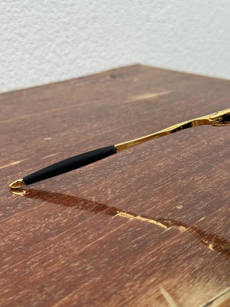 May include: Close-up of a gold-colored eyeglass arm with a black rubber grip. The arm is resting on a wooden surface. The end of the arm has a small gold loop.