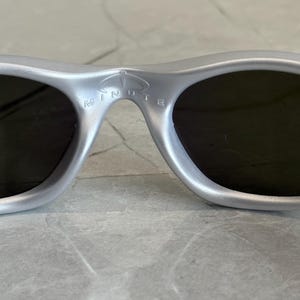 May include: Silver sunglasses with dark lenses. The bridge of the sunglasses features the word "MINUTE" in white lettering. The sunglasses have a sleek, modern design.