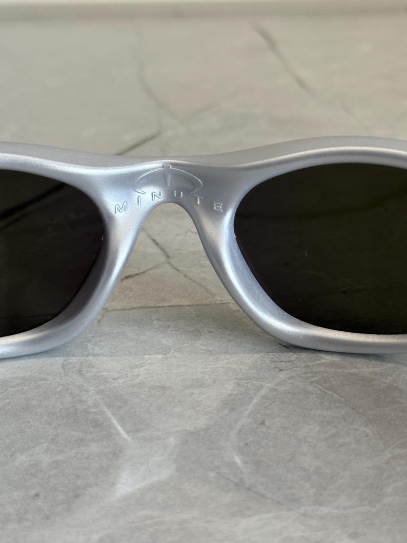 May include: Silver sunglasses with dark lenses. The bridge of the sunglasses features the word "MINUTE" in white lettering. The sunglasses have a sleek, modern design.