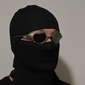 May include: Silver-framed, round sunglasses with dark lenses. The sunglasses are worn with a black balaclava. The balaclava covers the head and neck, leaving only the eyes visible.