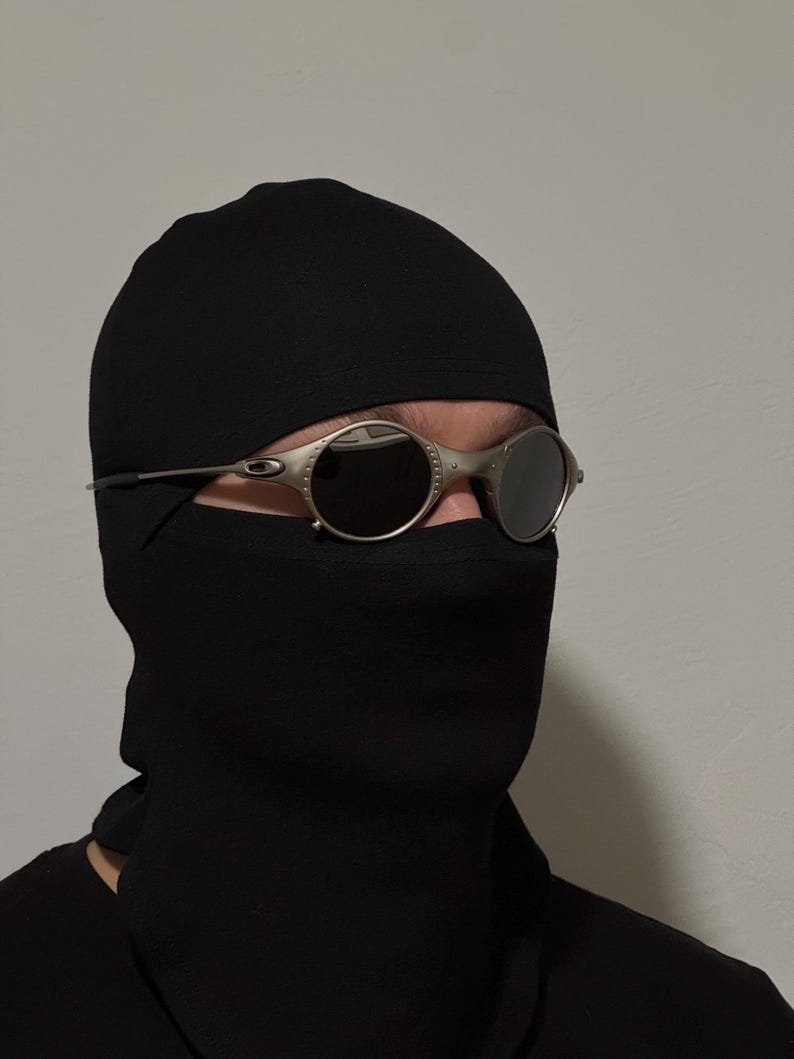 May include: Silver-framed, round sunglasses with dark lenses. The sunglasses are worn with a black balaclava. The balaclava covers the head and neck, leaving only the eyes visible.