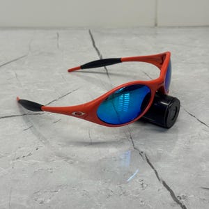 Op de afbeelding: Rode zonnebril met blauw gespiegelde glazen. De monturen zijn felrood, met zwarte accenten op de pootjes. De glazen reflecteren een helderblauwe tint. De zonnebril ligt op een zwart cilindrisch object.