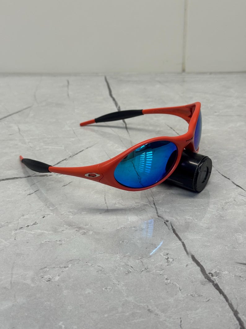 Op de afbeelding: Rode zonnebril met blauw gespiegelde glazen. De monturen zijn felrood, met zwarte accenten op de pootjes. De glazen reflecteren een helderblauwe tint. De zonnebril ligt op een zwart cilindrisch object.