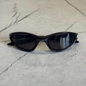 K&ouml;nnte beinhalten: Schwarze Sonnenbrille mit geschwungenem Rahmen und dunklen Gl&auml;sern. Der Markenname "Oakley" ist auf dem Rahmen aufgedruckt. Die Sonnenbrille liegt auf einer grauen Marmoroberfl&auml;che.
