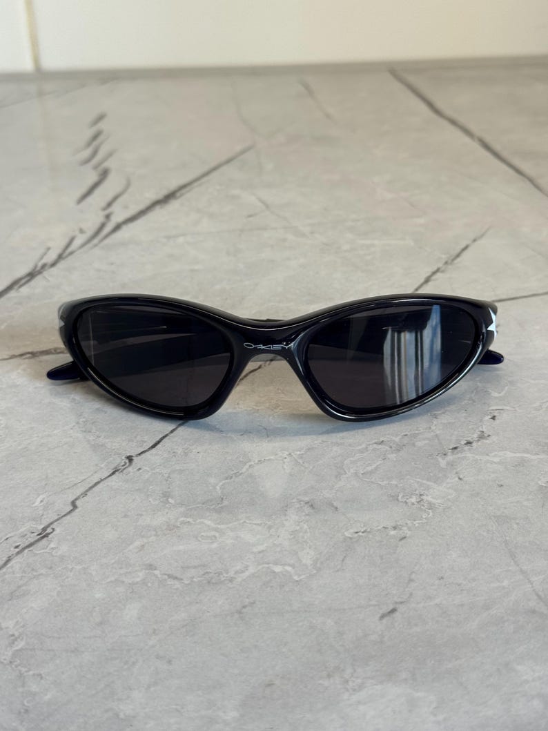 K&ouml;nnte beinhalten: Schwarze Sonnenbrille mit geschwungenem Rahmen und dunklen Gl&auml;sern. Der Markenname "Oakley" ist auf dem Rahmen aufgedruckt. Die Sonnenbrille liegt auf einer grauen Marmoroberfl&auml;che.
