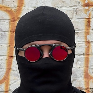 May include: Round, red-lensed sunglasses with a silver frame are worn over a black balaclava. The background is a weathered brick wall with orange paint.