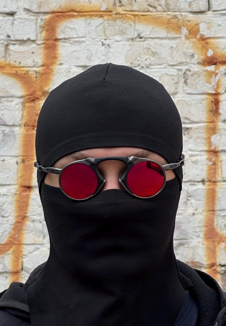 May include: Round, red-lensed sunglasses with a silver frame are worn over a black balaclava. The background is a weathered brick wall with orange paint.