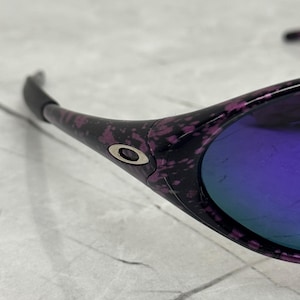 May include: Close-up of sunglasses with a black frame featuring a purple and pink speckled pattern. The lenses have a reflective purple hue. The Oakley logo is visible on the frame.