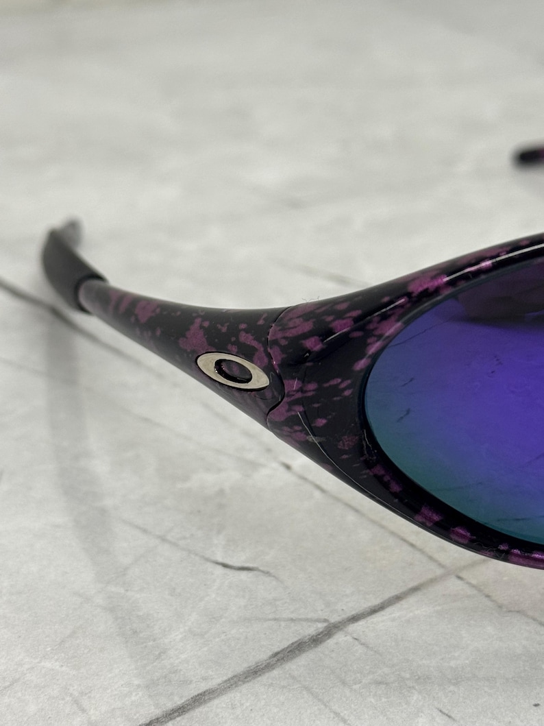 May include: Close-up of sunglasses with a black frame featuring a purple and pink speckled pattern. The lenses have a reflective purple hue. The Oakley logo is visible on the frame.