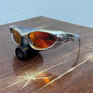 Puede incluir: Gafas de sol plateadas con lentes naranjas. Las gafas de sol tienen un dise&ntilde;o deportivo con una montura curvada y un peque&ntilde;o logotipo en la patilla. Las lentes son de color naranja vibrante y la montura es plateada.