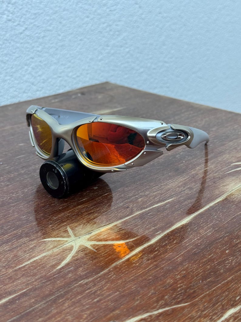 Puede incluir: Gafas de sol plateadas con lentes naranjas. Las gafas de sol tienen un dise&ntilde;o deportivo con una montura curvada y un peque&ntilde;o logotipo en la patilla. Las lentes son de color naranja vibrante y la montura es plateada.