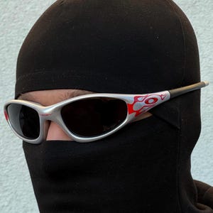 May include: Silver-framed sunglasses with red and white accents. The sunglasses have dark lenses and are worn with a black balaclava. The design on the frame is a flame-like pattern. The sunglasses are a sporty style.